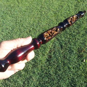 Puede incluir: Una varita m&aacute;gica de madera hecha a mano con un mango burdeos oscuro y una punta negra. El eje de la varita presenta una secci&oacute;n tallada llena de peque&ntilde;as piedras de color marr&oacute;n claro. La varita se sostiene sobre un fondo de hierba verde.