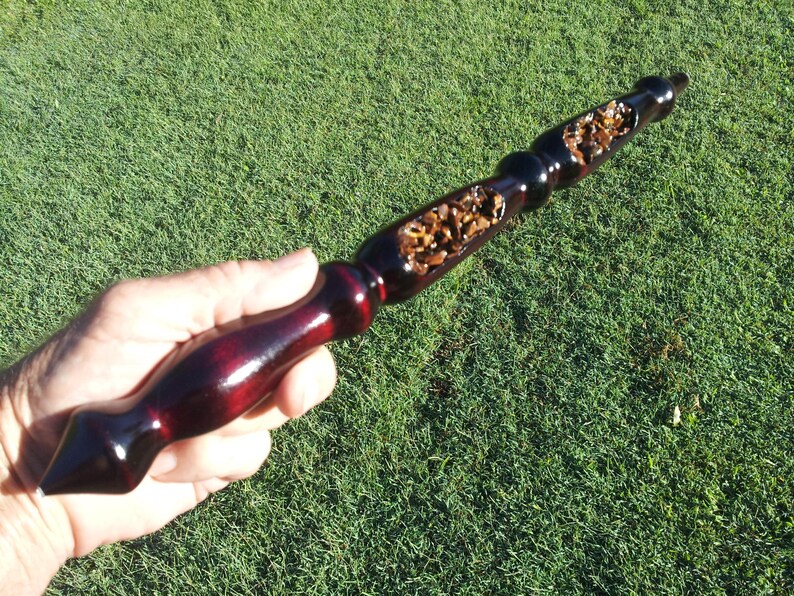 Puede incluir: Una varita m&aacute;gica de madera hecha a mano con un mango burdeos oscuro y una punta negra. El eje de la varita presenta una secci&oacute;n tallada llena de peque&ntilde;as piedras de color marr&oacute;n claro. La varita se sostiene sobre un fondo de hierba verde.