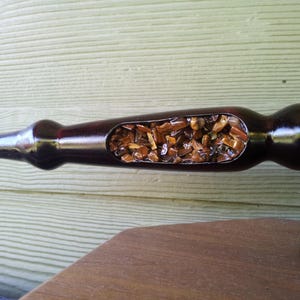 Puede incluir: Una pipa de madera marr&oacute;n oscuro con una secci&oacute;n ahuecada llena de peque&ntilde;as piedras de color &aacute;mbar. La pipa tiene un acabado liso y brillante y est&aacute; sobre una superficie de madera. Dise&ntilde;o &uacute;nico y artesanal.