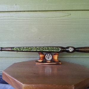 May include: A handcrafted wooden wand with a dark finish, inlaid with green stones. The wand features a pentagram design on the handle and is displayed on a wooden stand. The wand is approximately 14 inches long.