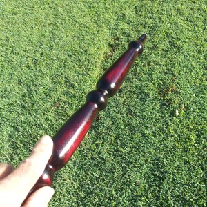 Puede incluir: Objeto de madera torneada, predominantemente rojo oscuro con detalles en negro. El art&iacute;culo tiene un acabado brillante y se coloca sobre hierba verde. El dise&ntilde;o del objeto es alargado, con una parte superior e inferior redondeadas.