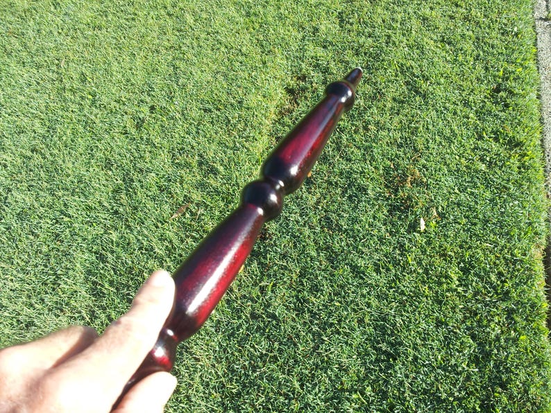 Puede incluir: Objeto de madera torneada, predominantemente rojo oscuro con detalles en negro. El art&iacute;culo tiene un acabado brillante y se coloca sobre hierba verde. El dise&ntilde;o del objeto es alargado, con una parte superior e inferior redondeadas.