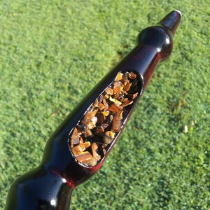 Puede incluir: Una pipa de fumar burdeos oscuro con un tallo largo y curvado. La pipa tiene una cazoleta grande, de forma ovalada, llena de tabaco marr&oacute;n. El fondo es un c&eacute;sped verde borroso.