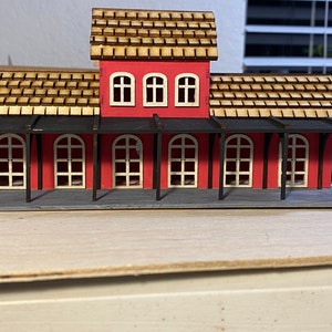 May include: A red wooden model train station with a black roof and a gray platform. The station has four windows and a small awning.