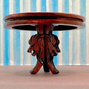 May include: A dark brown wooden table with a round top and a base carved with two lion heads. The table is on a striped blue and white background.