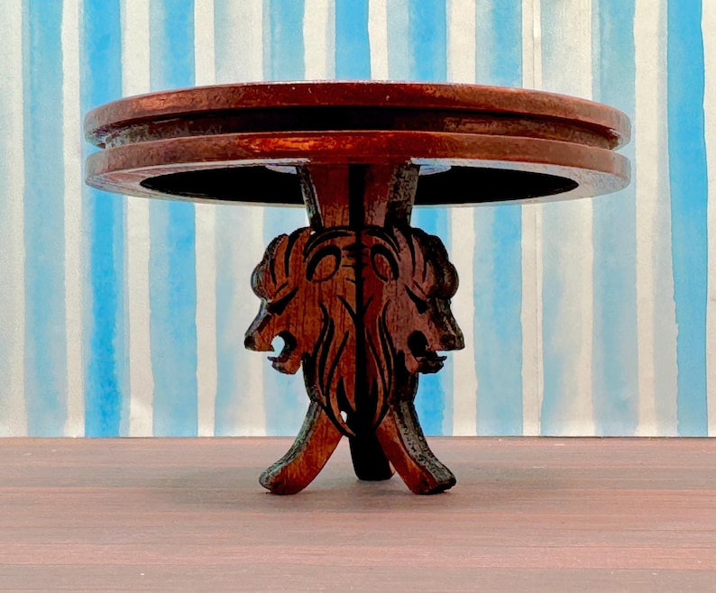 May include: A dark brown wooden table with a round top and a base carved with two lion heads. The table is on a striped blue and white background.