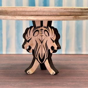 May include: A wooden table with a round top and a base carved in the shape of three lion heads. The table is made of light brown wood and has a natural finish.