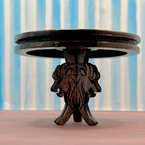 May include: A dark brown wooden table with a round top and a carved base featuring three stylized animal heads. The table is on a wooden surface.