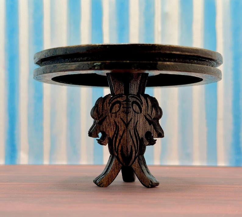 May include: A dark brown wooden table with a round top and a carved base featuring three stylized animal heads. The table is on a wooden surface.