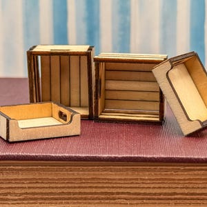May include: Four miniature wooden crates and trays. The crates have a light brown color with dark brown trim. Two crates are open, and two are trays. The crates have a slatted design. The trays have cut-out handles. The items are on a maroon surface.