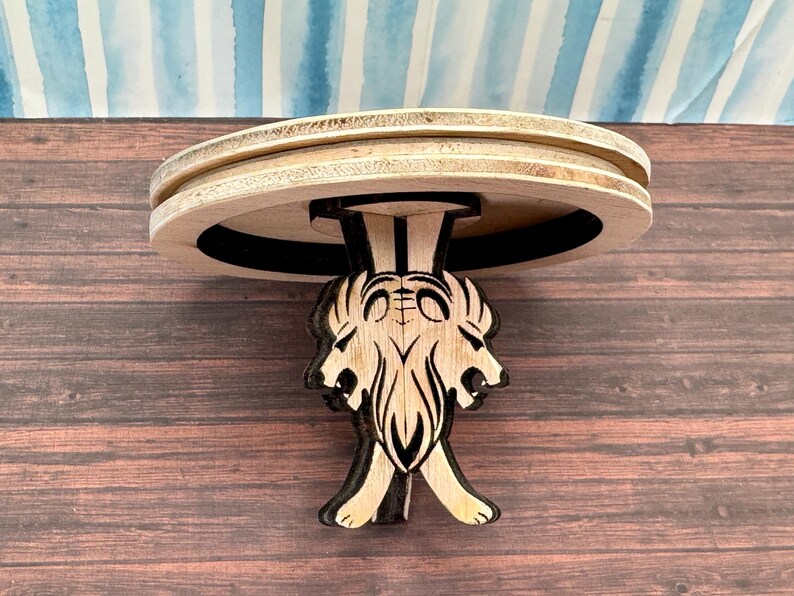 May include: A wooden table with a round top and three legs shaped like lion heads. The table is made of light wood and has a dark brown stain.