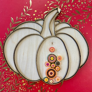 May include: A white wooden pumpkin with a brown stem and a decorative design of colorful paper circles. The pumpkin is on a red background with gold glitter.