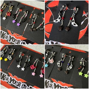 May include: A selection of handmade earrings with diverse designs, such as guitars, stars, and abstract shapes. The earrings incorporate silver-tone metal chains and vibrant accents in red, pink, and yellow. Each pair is presented on a black card with the text "KE Your D".