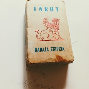 May include: A vintage tarot card deck box with the word "TAROT" in blue letters at the top. Below, the words "BARAJA EGIPCIA" are printed in blue. A red sphinx illustration is featured on the box. The box is slightly worn.