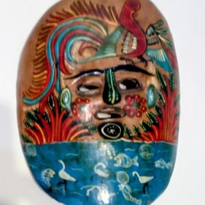 May include: Hand-painted, oval-shaped mask with a brown background and a blue lower section. The mask features a face with a green nose, and a red bird. The blue section depicts water with white birds and fish.
