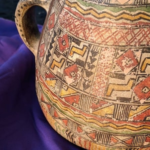 May include: A large, brown ceramic jug with a geometric pattern in red, yellow, green, and black. The jug has a wide mouth and a handle.