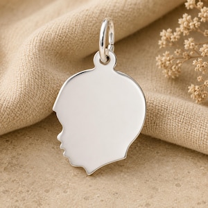 May include: A polished silver pendant in the shape of a child's profile. The pendant has a smooth, reflective surface and a small loop for attaching to a chain. The design is simple and elegant, suitable for a necklace or bracelet.