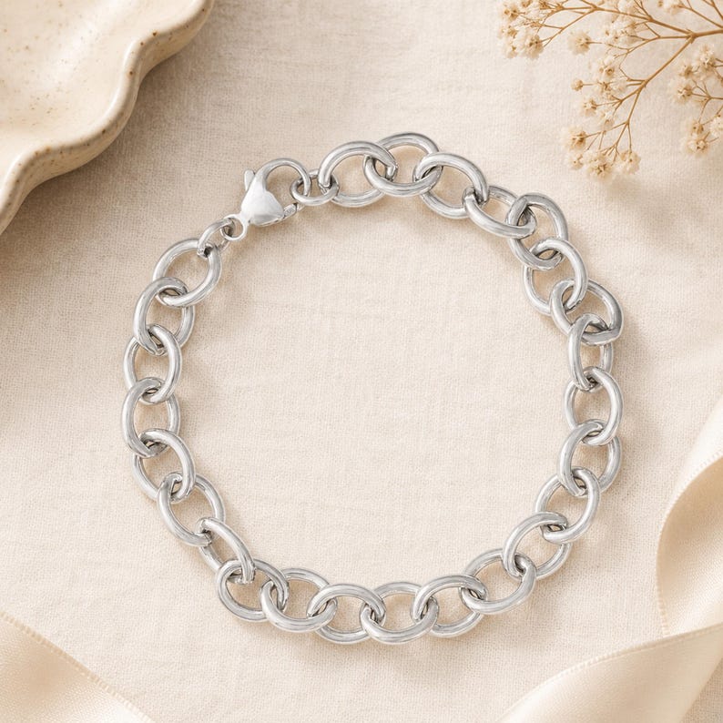 Op de afbeelding: Een zilverkleurige kettingarmband met grote ovale schakels. De armband is gemaakt van glanzend metaal en heeft een karabijnsluiting. De sieraden worden gepresenteerd op een neutrale ondergrond met decoratieve elementen.