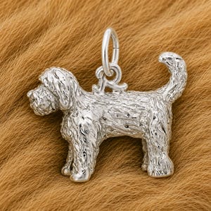 May include: A polished silver charm in the shape of a dog with a textured, shaggy coat. The dog is standing in profile with its head turned slightly toward the viewer. A circular silver jump ring is attached to the top of the dog's back.