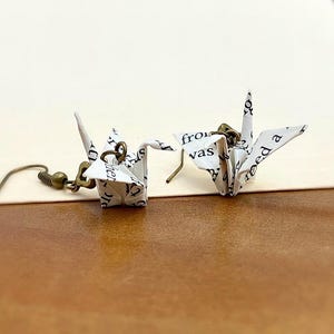 May include: A pair of origami crane earrings made from white paper with black text. The cranes are hanging from bronze hooks.