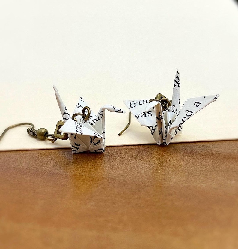 May include: A pair of origami crane earrings made from white paper with black text. The cranes are hanging from bronze hooks.