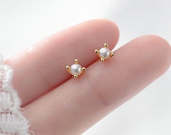 May include: A pair of small, gold-colored stud earrings, each featuring a central white pearl. The earrings have a four-point design, with the pearl at the center of each point. The earrings are displayed on a person's finger.