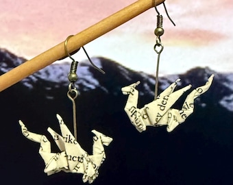 May include: A pair of handmade origami dragon earrings. The dragons are crafted from paper printed with text, featuring a white and cream color scheme. The earrings hang from silver-colored hooks, with a wooden dowel in the background.