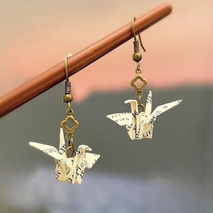 May include: A pair of earrings featuring origami cranes made from white paper with black text. The cranes are suspended from gold-toned chains with a small, decorative charm.