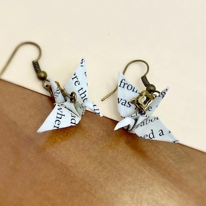 May include: A pair of origami paper crane earrings with bronze hooks. The cranes are made from white paper with black text printed on them.