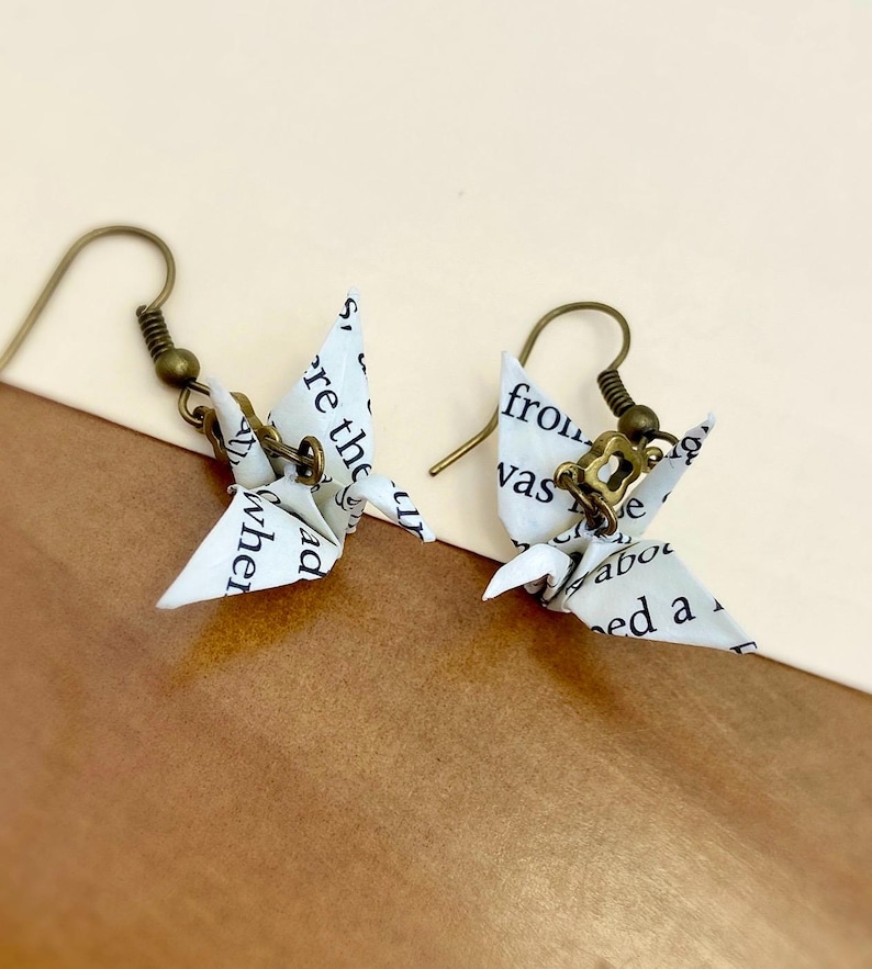 May include: A pair of origami paper crane earrings with bronze hooks. The cranes are made from white paper with black text printed on them.