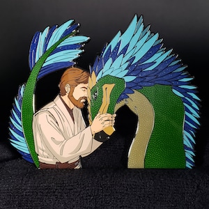 May include: A green and blue enamel pin featuring a man with a beard and a green and blue dragon. The man is wearing a white robe and the dragon has a long neck and a blue and green feathered crest.