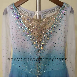 May include: A light blue figure skating dress with long sleeves and a V-neckline. The dress is decorated with iridescent rhinestones and has a gradient design. The text "etsy: customicedress" is visible at the bottom of the dress.
