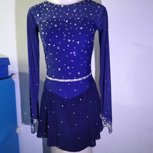 Puede incluir: Un vestido de patinaje art&iacute;stico azul oscuro con mangas largas y un dise&ntilde;o brillante. El vestido tiene un cintur&oacute;n plateado y una falda con un escote en V.