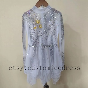 May include: White figure skating dress with long sleeves and a sparkly design. The dress has a high neck and a layered skirt. The dress is decorated with silver and gold rhinestones. The dress is on a hanger. etsy:customicedress