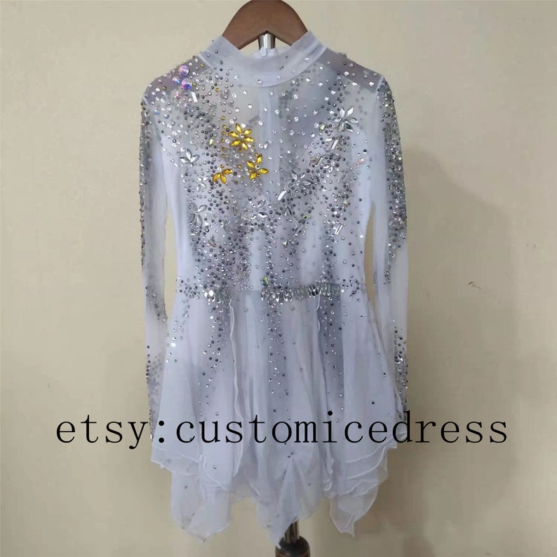 May include: White figure skating dress with long sleeves and a sparkly design. The dress has a high neck and a layered skirt. The dress is decorated with silver and gold rhinestones. The dress is on a hanger. etsy:customicedress