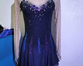 May include: A navy blue figure skating dress with a sheer neckline and long sleeves. The bodice is embellished with sparkling rhinestones and crystals in shades of blue and purple. The skirt is a flowing, layered design.