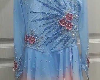 Op de afbeelding: Een lichtblauwe kunstschaatsjurk met lange mouwen en een roze ombre rok. De jurk is versierd met roze bloemen en strass.