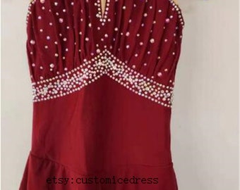 Puede incluir: Vestido de patinaje artístico sin mangas de color burdeos con un escote de ojo de cerradura y un dobladillo festoneado. El vestido está adornado con pedrería.