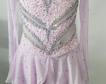 Puede incluir: Un vestido de patinaje artístico de color lavanda con mangas largas y un corpiño de encaje adornado con pedrería. La falda es de capas y tiene un dobladillo festoneado.
