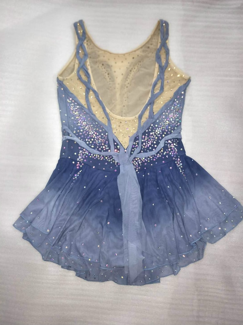 May include: A blue and white figure skating dress with a mesh bodice and a layered skirt. The dress is decorated with rhinestones and has a criss-cross back design.