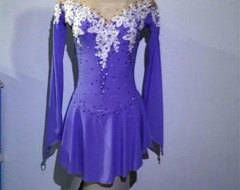 Puede incluir: Un vestido de patinaje artístico morado con mangas largas y un corpiño de encaje blanco. El vestido tiene un escote de malla transparente y está decorado con pedrería.