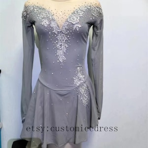 Könnte beinhalten: Ein graues Eiskunstlaufkleid mit langen Ärmeln und einem weißen Spitzenmuster. Das Kleid hat einen Mesh-Ausschnitt und einen leicht ausgestellten Rock. Das Kleid ist mit Strasssteinen verziert.