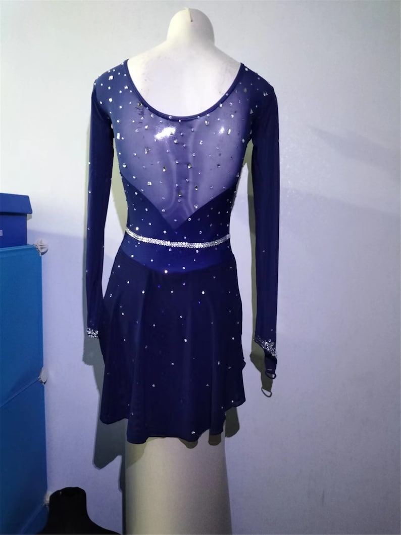 Puede incluir: Un vestido de patinaje art&iacute;stico azul marino con mangas largas y un panel trasero de malla transparente. El vestido est&aacute; decorado con diamantes de imitaci&oacute;n y un cintur&oacute;n de diamantes de imitaci&oacute;n plateado.
