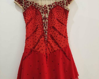 Puede incluir: Un vestido de patinaje artístico rojo con un escote nude transparente y mangas cortas. El corpiño está muy adornado con pedrería y cristales rojos, con una falda acampanada. El vestido está colgado de una percha azul contra una pared blanca.