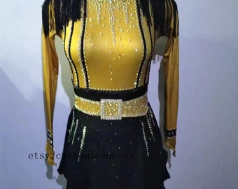 May include: A gold and black figure skating dress with long sleeves, a black belt with a gold buckle, and a black skirt with rhinestones. The dress has black fringe on the shoulders.