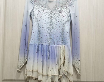 Op de afbeelding: Een lichtblauwe kunstschaatsjurk met een wit ombre-effect en een hoge halslijn. De jurk is bedekt met strass en heeft een gelaagde rok.