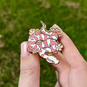 May include: A pink and white snake with a gold outline is curled up with pink flowers. The snake is a pin with a gold back.