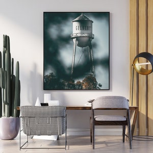 May include: A black and white photograph of a water tower with the word "GILBERT" on it. The water tower is set against a cloudy sky. The photograph is framed and hanging on a white wall in a room with a wooden desk and a chair.