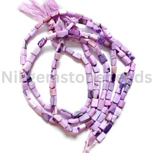 Puede incluir: Hebras de cuentas rectangulares moradas con una borla. Las cuentas tienen un aspecto veteado con tonos lavanda, lila y morado más oscuro. La borla es de color morado claro a juego. Las cuentas están ensartadas.