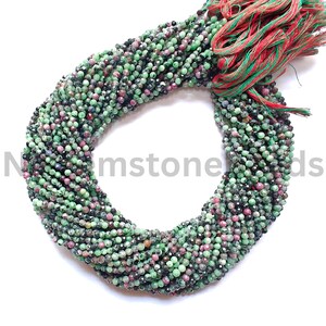 May include: Strands of faceted gemstone beads in shades of green, pink, and black. The beads are strung together, forming a circular shape. The ends of the strands are tied with red and green threads. The beads are small and appear to be semi-precious stones.